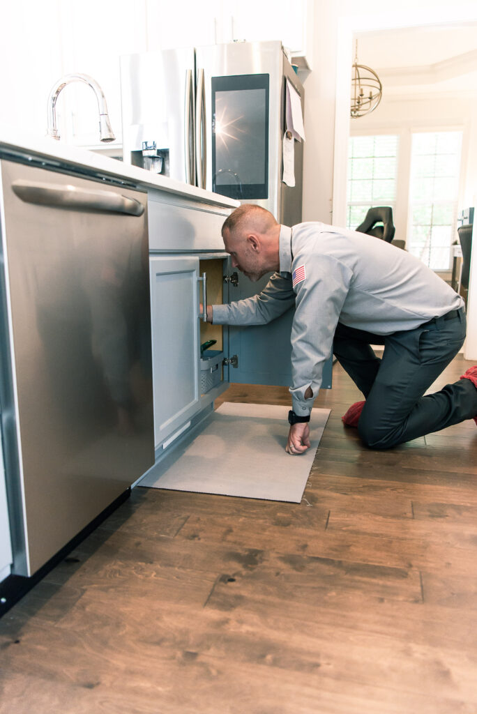 Plumber inspects under-sink plumbing in a kitchen with wood floors.