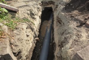 A sewer drain pipe is being installed in a narrow trench dug through the ground, with the black pipe surrounded by excavated soil.